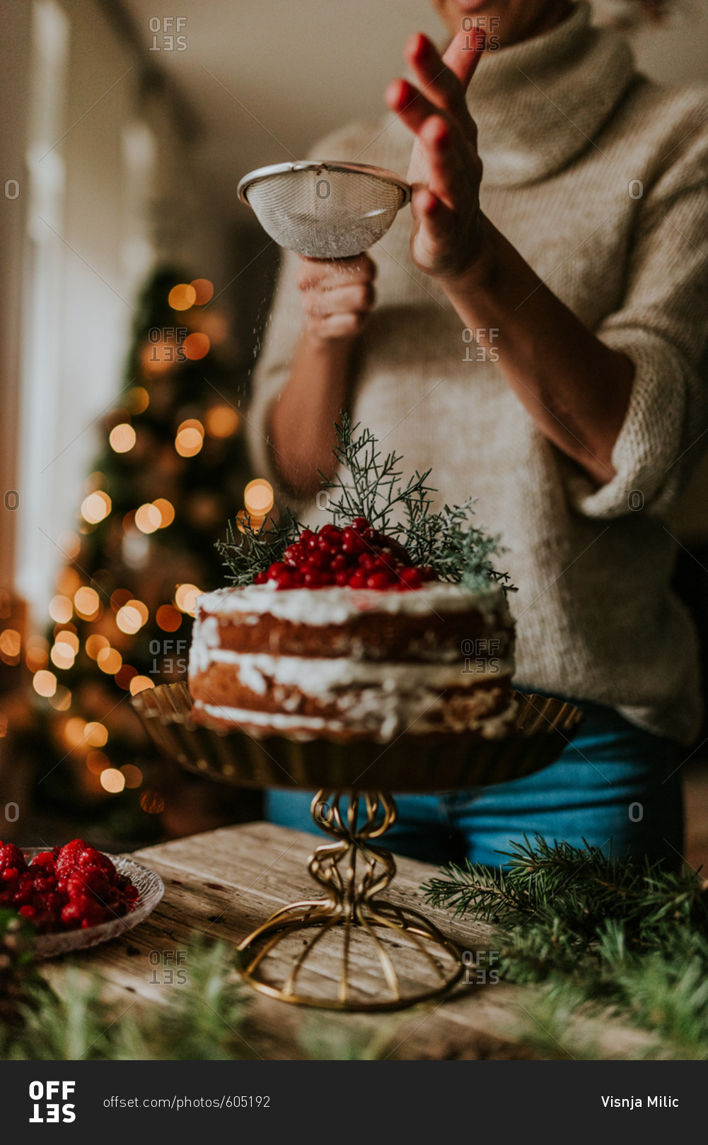Woman decorating winter inspired naked cake with powdered sugar with Christmas tree in the background