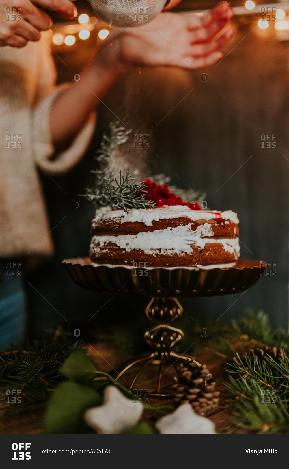 Woman decorating winter inspired naked cake with powdered sugar with Christmas lights in the background