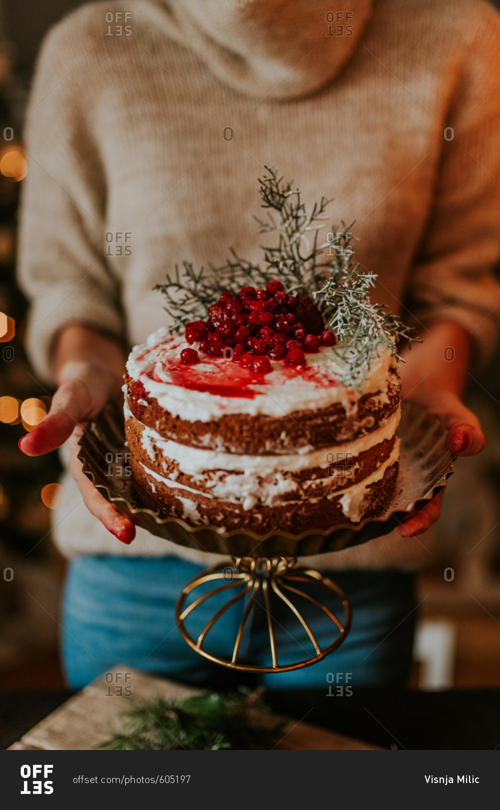 Woman holding naked cake on a gold pedestal decorated with strawberries and redcurrant with Christmas lights in the background