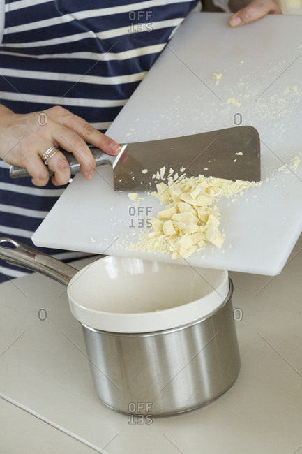 Close up of person wearing a blue and white stripy apron holding a chopping board and cleaver, scraping chopped white chocolate into a white bowl over a metal pot