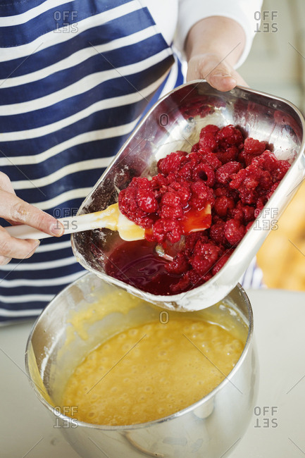 Close up high angle view of person wearing a blue and white stripy apron holding a metal dish, adding fresh raspberries to cake batter in a metal mixing bowl