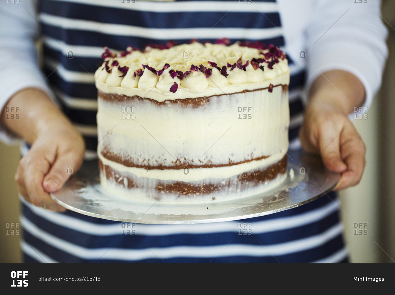 Close up of person wearing a blue and white stripy apron holding a cake decorated with cream and purple flower petals