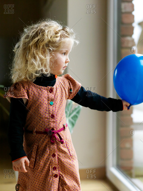 A portrait of a smiling young blonde girl looking through the window holding a blue birthday balloon at home with a short depth of field and blurred background