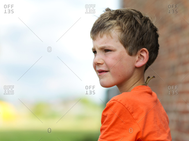 An outdoor portrait of a young boy looking ahead, near a house brick wall on a blurry garden background, green and light blue, short depth of field