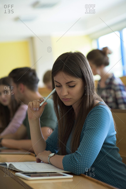 Young female using tablet computer while studying at desk with classmates at desk in classroom