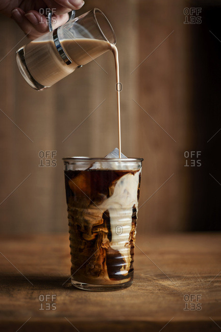 Glass of iced coffee with vintage creamer pouring into the glass on warm wood tones