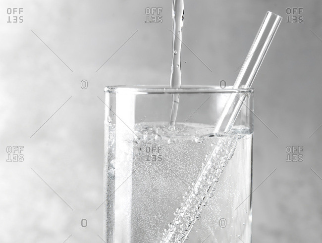 Detail of sparkling water being poured into a clear glass with a clear straw on a gray metal background