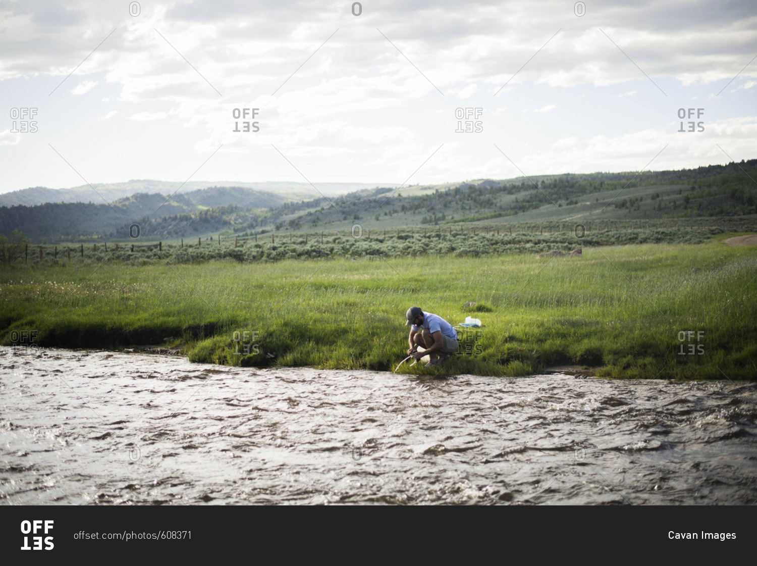 Hiker filling water bottle while crouching by river against cloudy sky