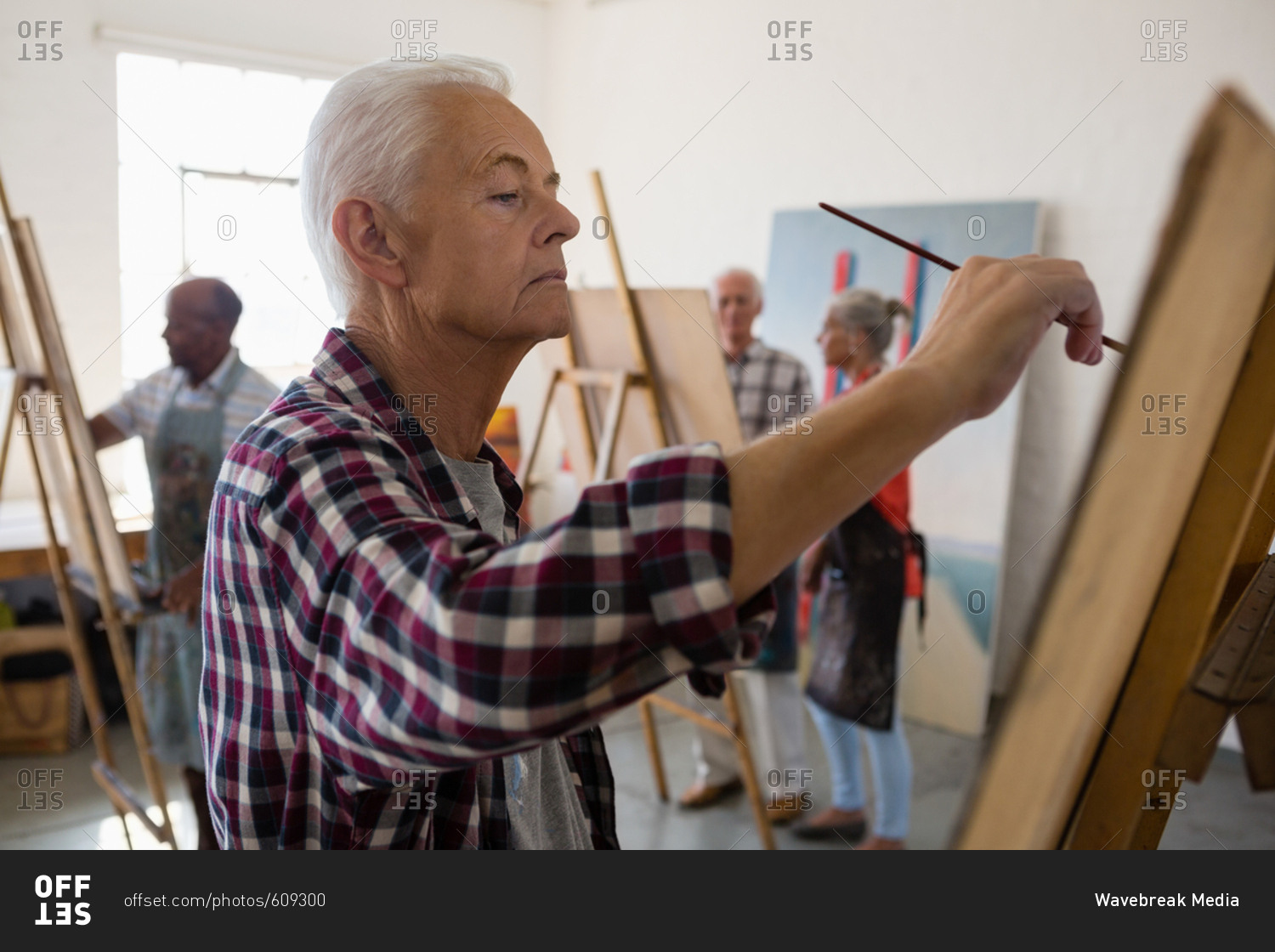 Side view of senior man painting with friends in background at art class
