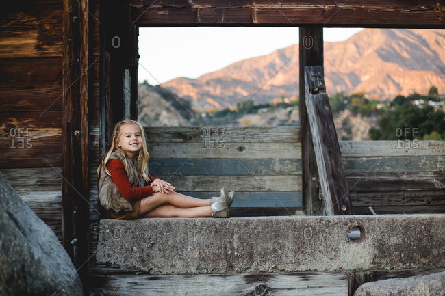 A well dressed young girl sitting on a rustic concrete and wood wall smiling
