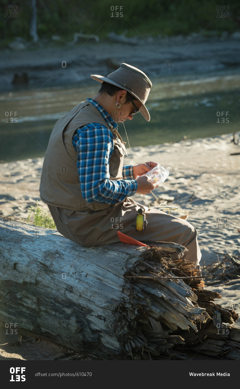 Side view of man looking at fishing bait while sitting on fallen tree trunk during sunny day