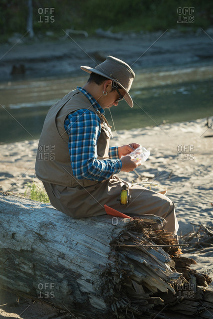 Side view of man looking at fishing bait while sitting on fallen tree trunk during sunny day