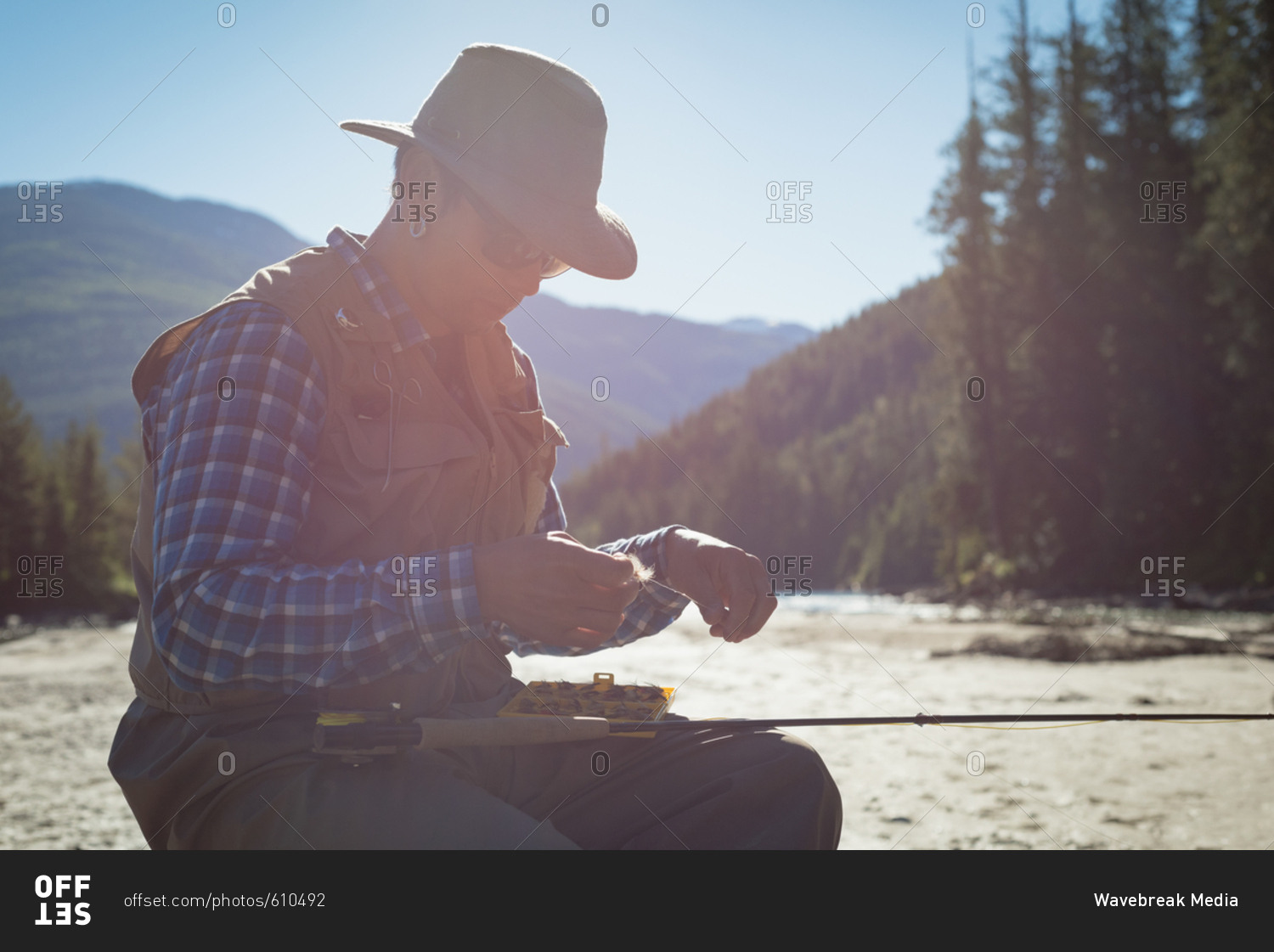 Young man tying fishing hook on rod while sitting at riverbank during sunny day