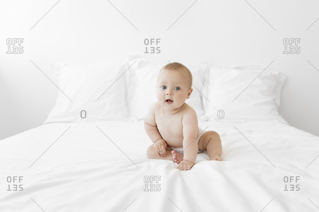 Baby boy sitting on a bed with white bedding, looking towards the camera