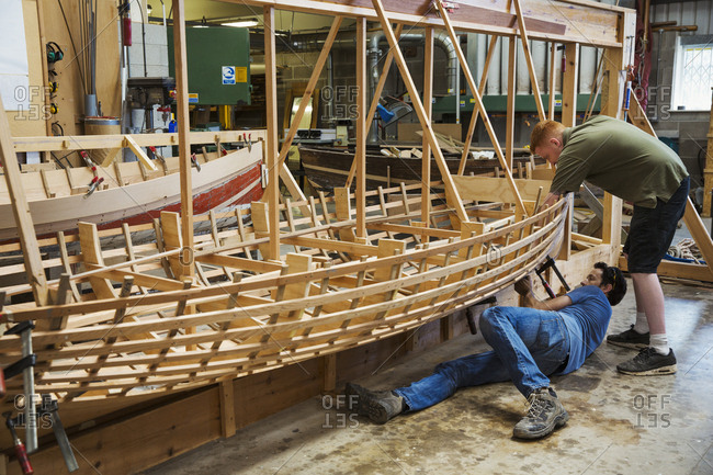 Two men in a boat-builder\'s workshop, working together on a wooden boat hull