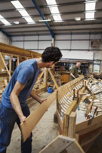Two men in a boat-builder\'s workshop, working together on a wooden boat hull