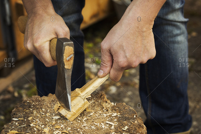 Close up of a man's hand holding an axe, cutting and shaping a small piece of wood on a splitting block covered in wood shavings