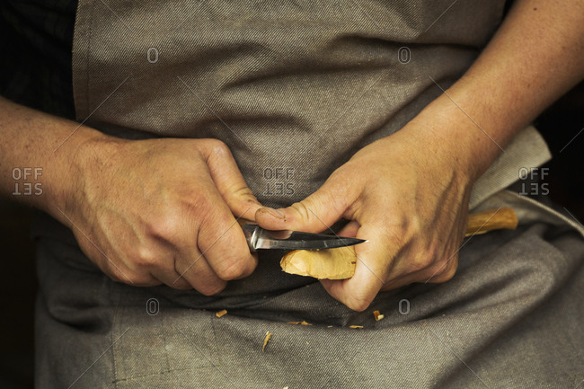Close up of a craftsman's hands pressing and shaping a small piece of wood into a spoon with a sharp knife blade, shaping the bowl back