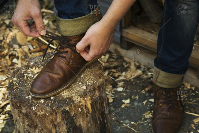 A man in denim jeans with turnups sitting leaning down to tie his shoe laces of his brown leather shoes, one foot up on a splitting block