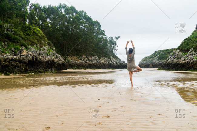 Back view of woman standing in yoga pose on one leg on sandy bay in ocean.