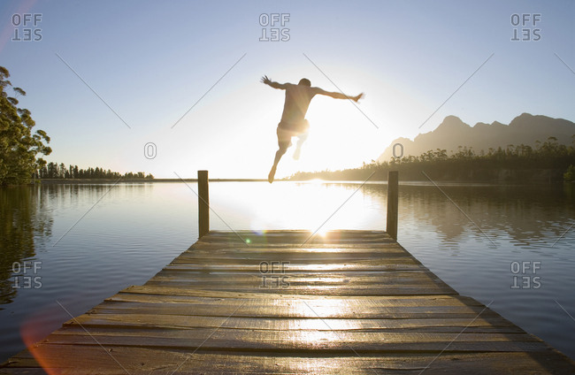 Man jumping from jetty into lake at sunset, rear view (lens flare, backlit)