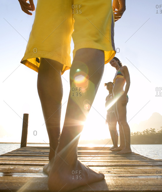 Family standing on jetty, man in yellow swimming shorts preparing to jump into lake, low-section, rear view (surface level, lens flare)
