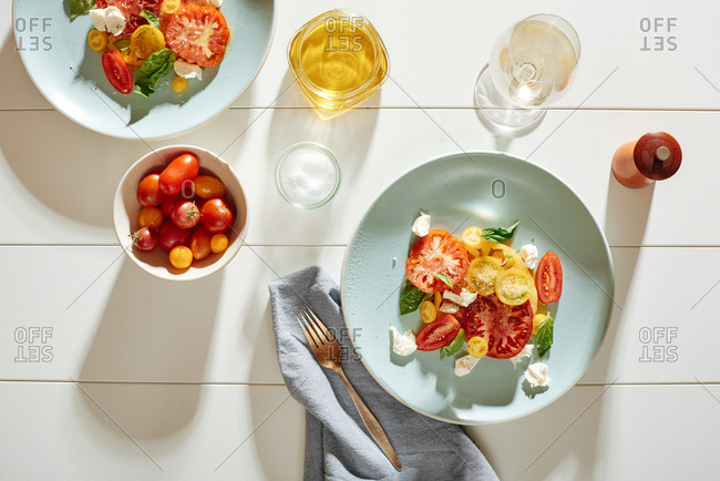 Heirloom tomato caprese salad with fresh basil leaves, mozzarella with kosher salt and fresh ground pepper.  On the table are a small bowl of cherry tomatoes, kosher salt and olive oil jar and a glass of white wine