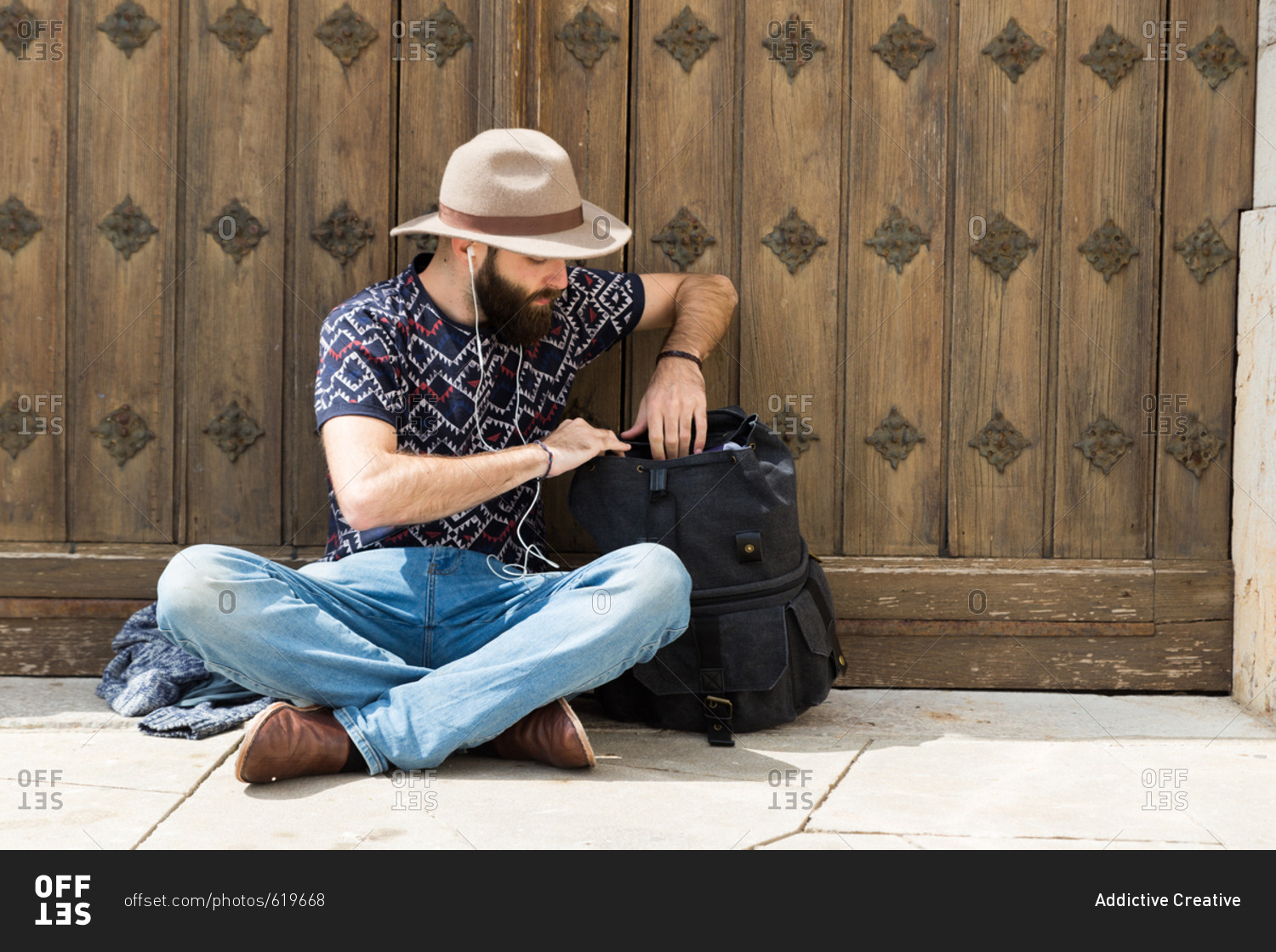 Horizontal outdoors shot of a man sitting on the ground and searching in a backpack while listening to music