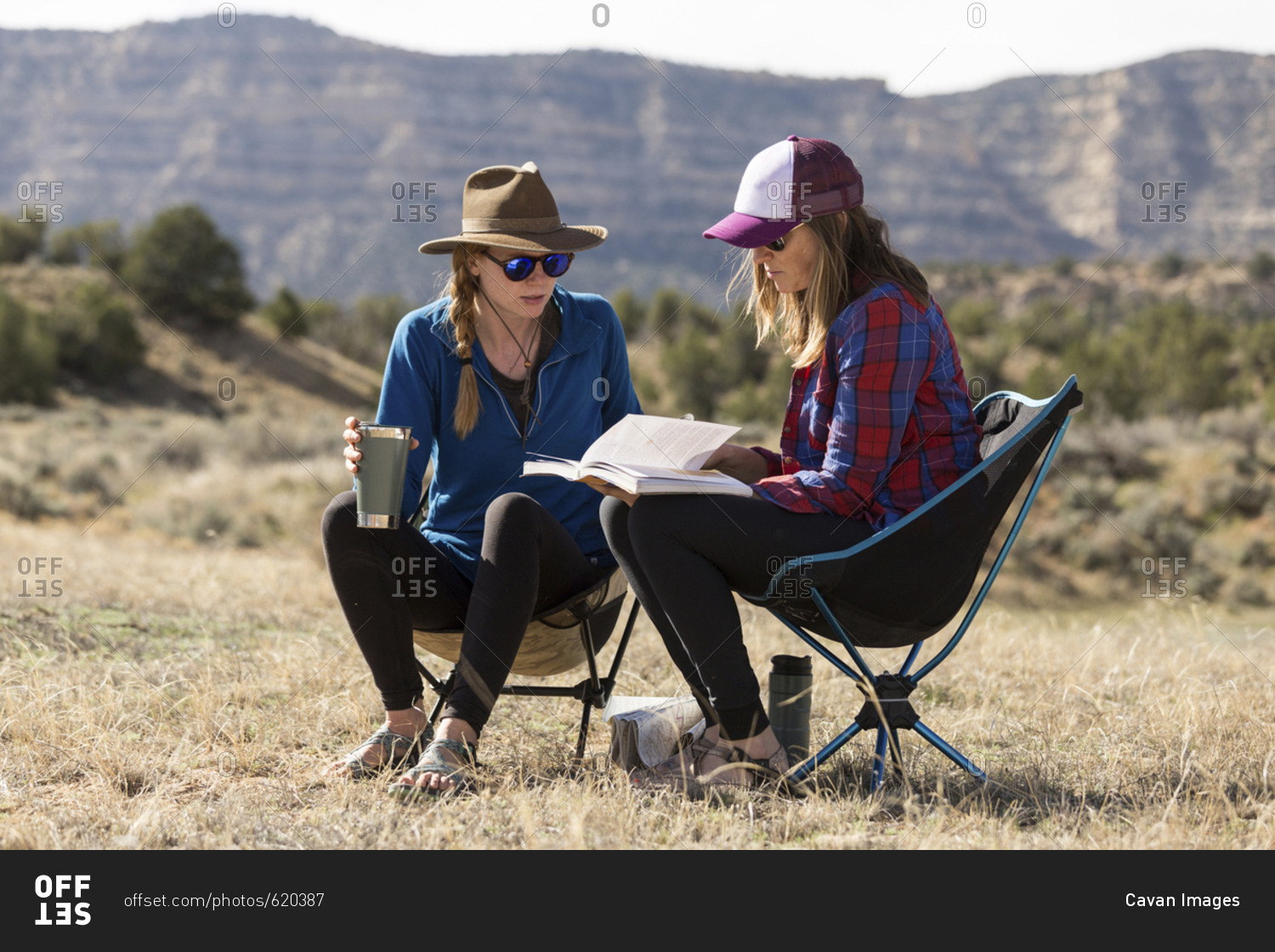 Female friends looking at book while sitting on camping chairs at field against mountains during sunny day