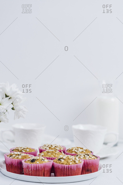 Pumpkin oatmeal muffins served on a white plate photographed on a white background. White flowers, white cups and a bottle of milk accompany.