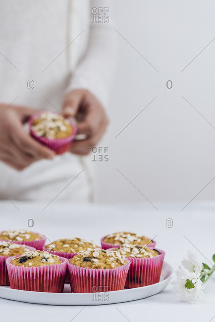 A woman in white clothes removing a pumpkin muffin from a pink muffin liner photographed from front view. A plate full of muffins and white flowers on the table accompany.