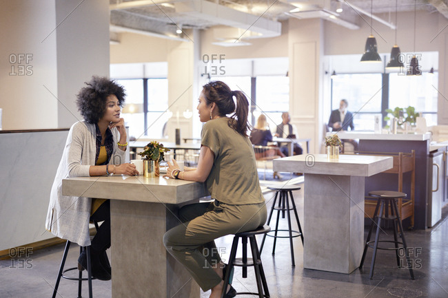 Businesswomen talking while sitting in cafeteria at creative office with colleagues in background