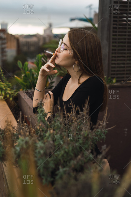 Pretty brunette posing sensually while smoking on balcony with cityscape on background