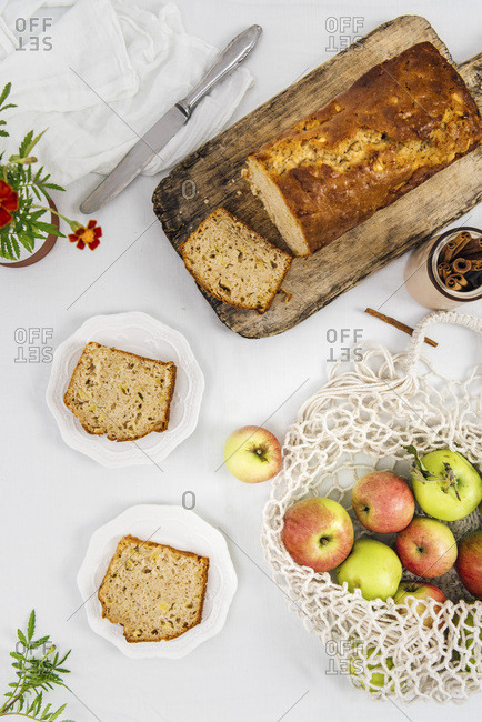 Cinnamon apple bread sliced on a wooden cutting board photographed from top view. Apples in a net bag and apple bread slices on two plates accompany.