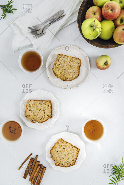 Cinnamon apple bread slices served on three white plates photographed from top view. Three cups of tea and apples in a wooden bowl accompany.