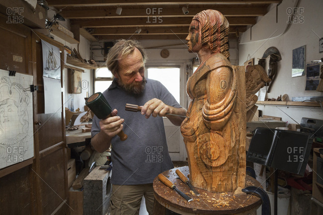 A wood carver standing in his workshop, using hand tools to shape and create decoration on a work in progress, a wooden female ship's figurehead.