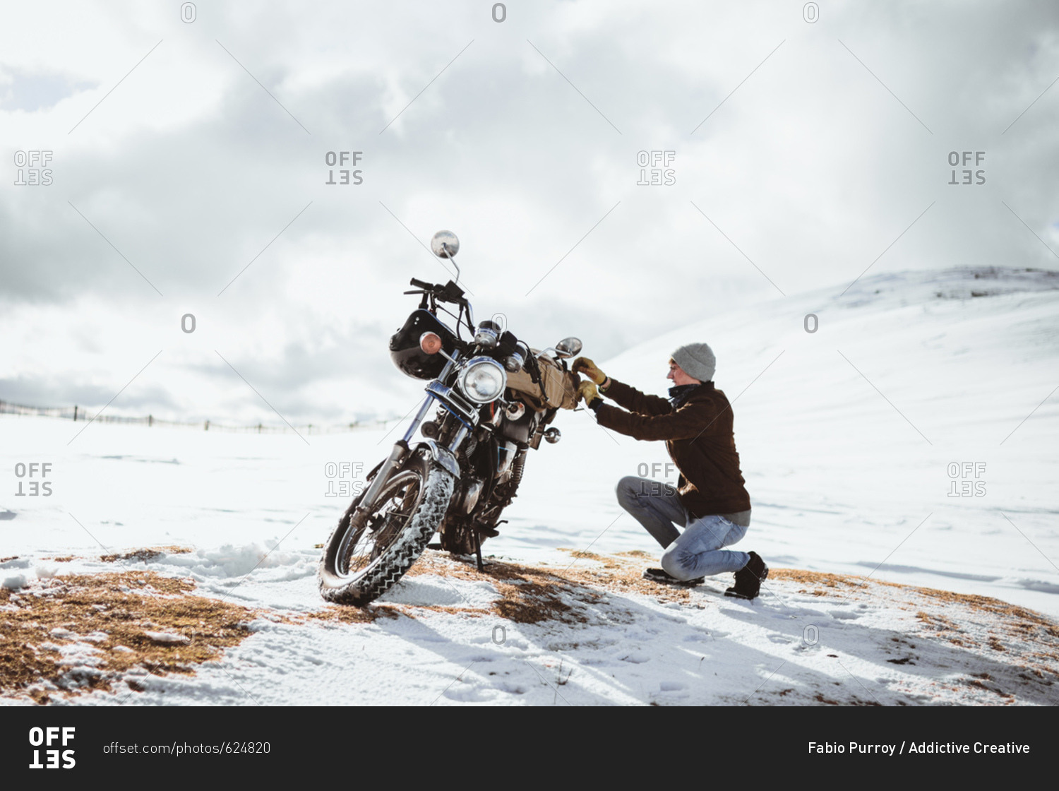 Side view of man arranging motorcycle while traveling alone across snowy mountains valley.