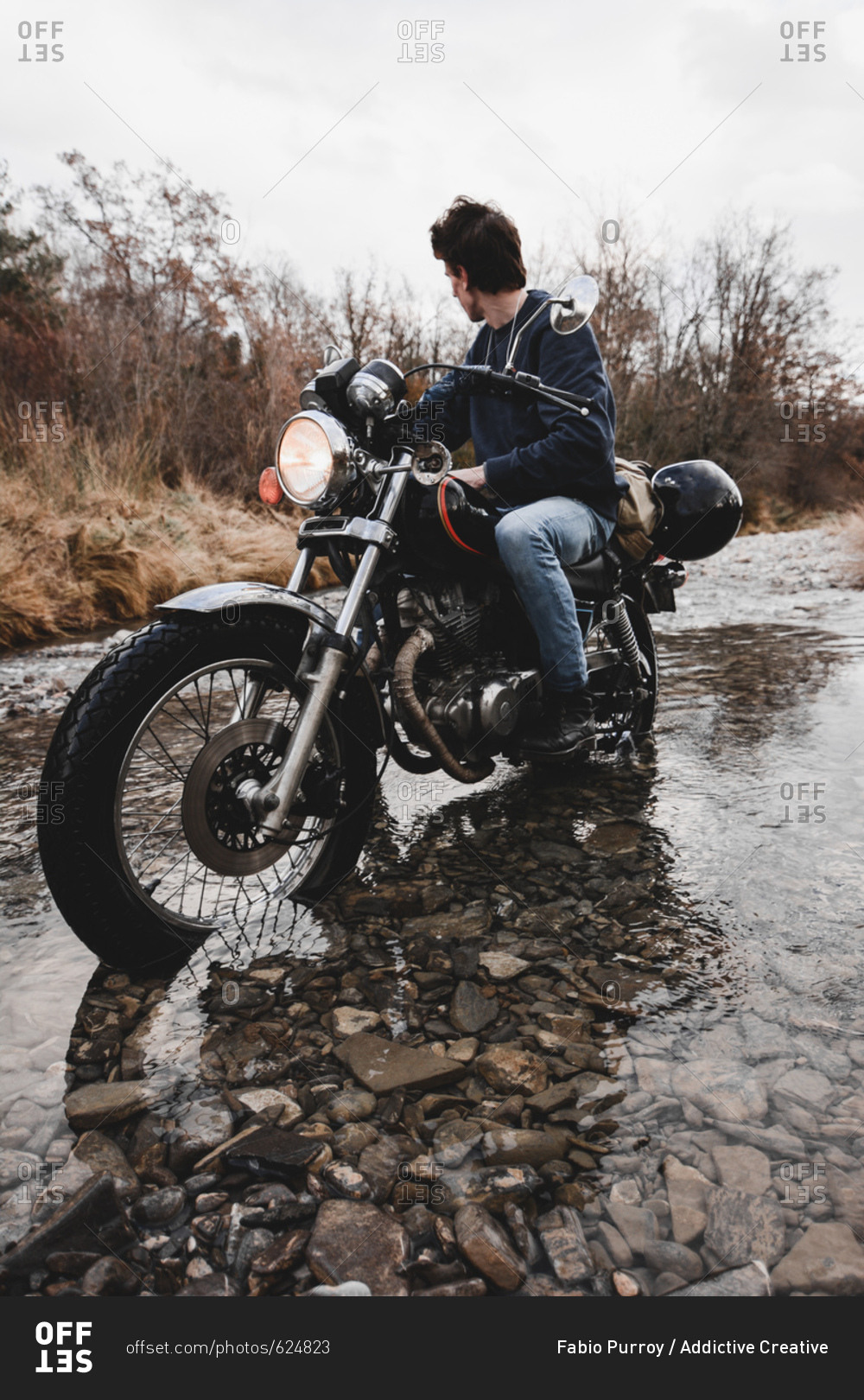 Anonymous man on motorcycle riding through water of creek in autumnal mountains while traveling alone.