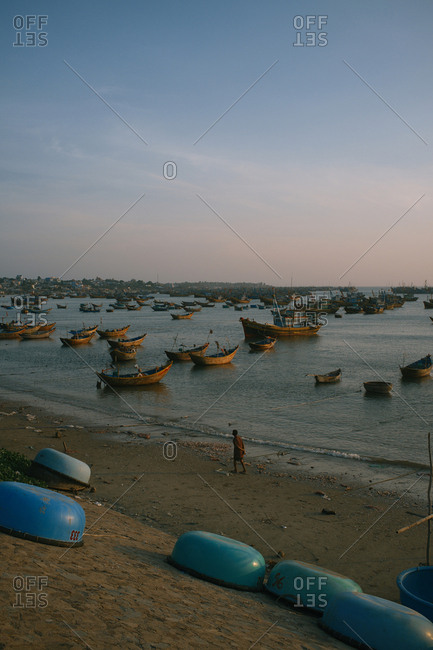 Panoramic view on traditional fishing boats in Mui Ne harbor, Phan Thiet city, Vietnam. Quiet water surface with small ripples, cloudless sky at sunset. Silhouette of local man walking along seashore and enjoying amazing view.