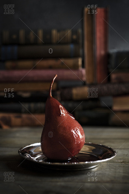 A wine poached pear on silver tray on a table with anitque books in the background. Rustic & moody feel.