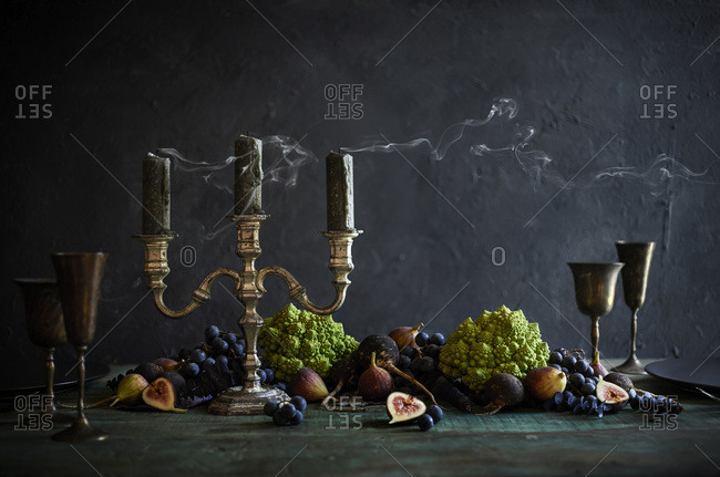 A gorgeous, creepy halloween tablescape with rustic candelabra with blown out candles. Vegetable spread across table with romanesco broccoli, concord grapes, figs and black radishes.