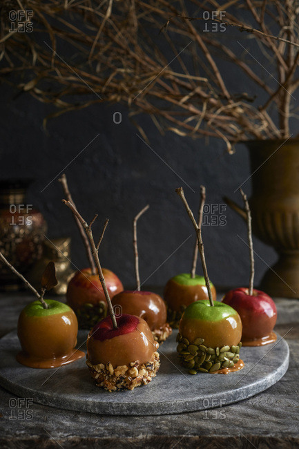 Marble tray full of caramel apples with stick handles covered in nuts and seeds. Moody fall foliage in the background.