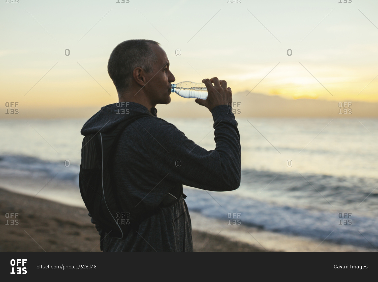 Side view of man drinking water while standing at beach during sunset