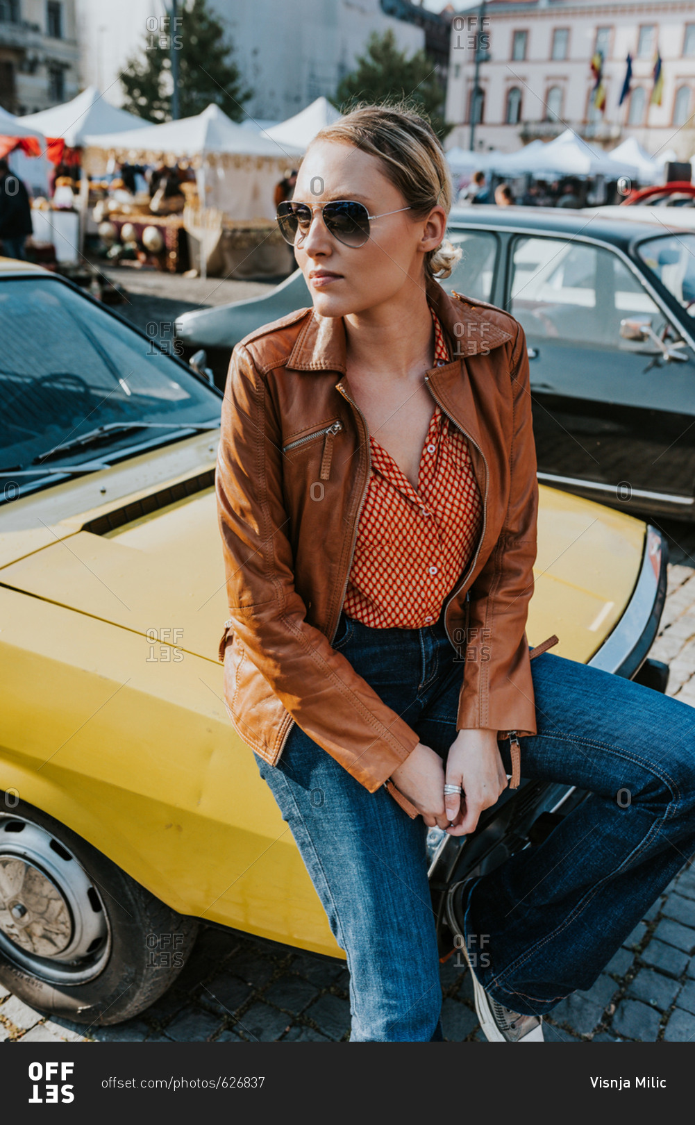 Blonde woman in late twenties sitting on yellow oldtimer car wearing retro seventies clothes style and aviator glasses