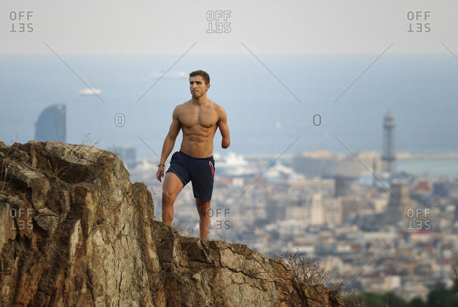 Physically challenged shirtless man looking away while standing on cliff against cityscape