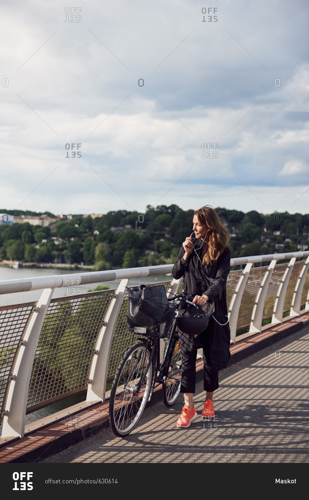 Mature woman talking through headphones while walking with bicycle on footbridge against sky