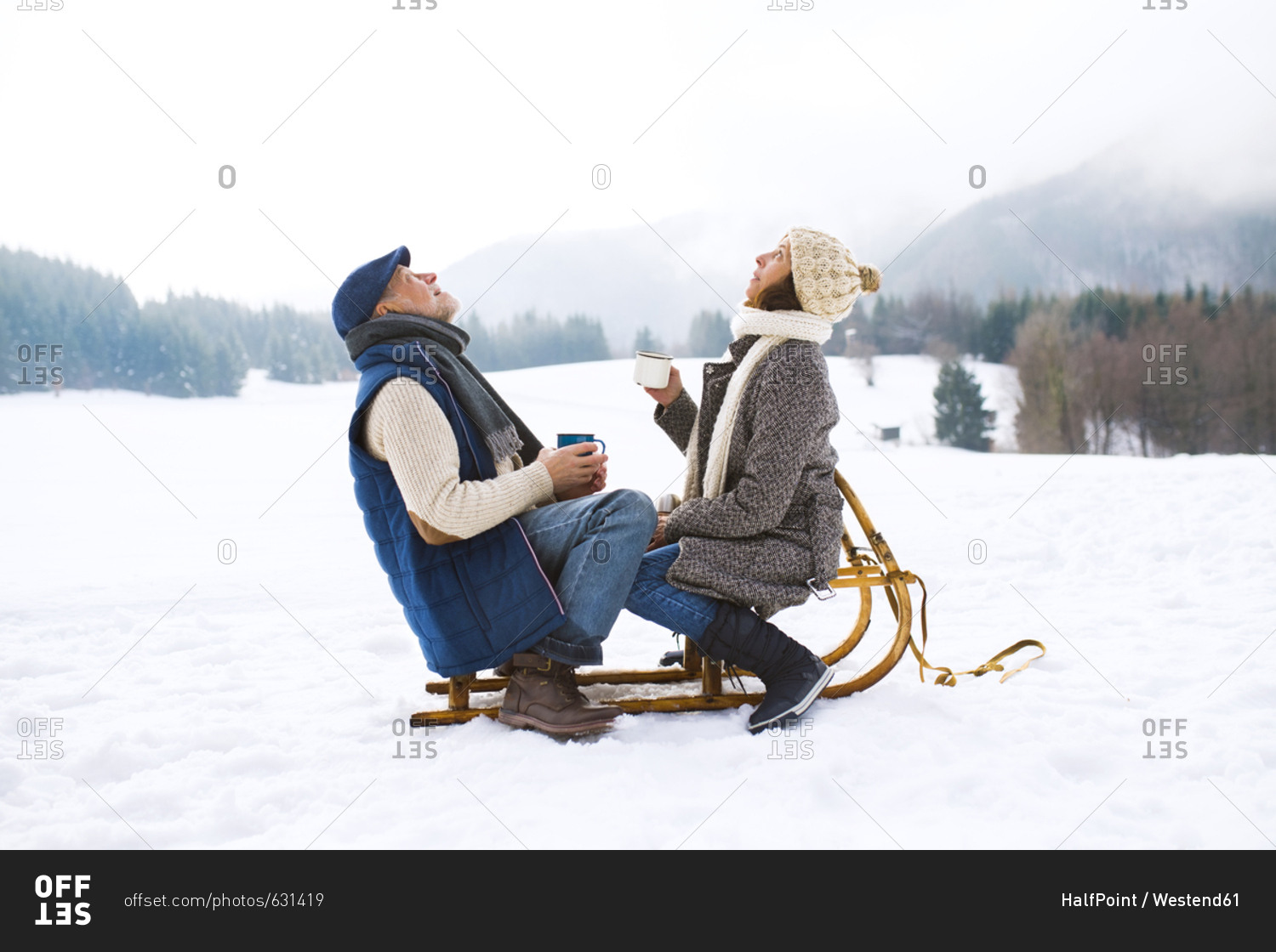 Senior couple sitting on sledge with hot beverages in snow-covered winter landscape looking up