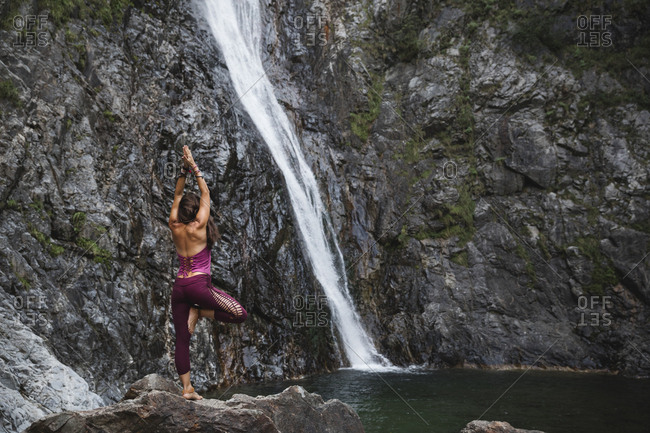 Italy- Lecco- woman doing Tree Yoga Pose on a rock near a waterfall