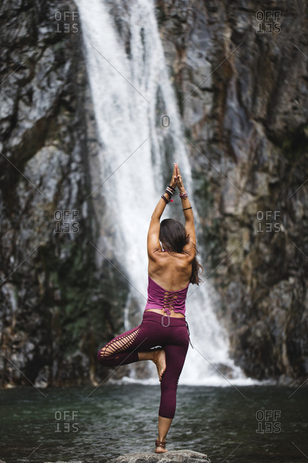 Italy- Lecco- woman doing Tree Yoga Pose on a rock near a waterfall