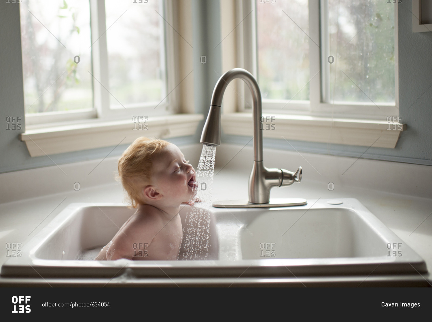 Cute baby boy sticking out tongue under running water from faucet in kitchen sink